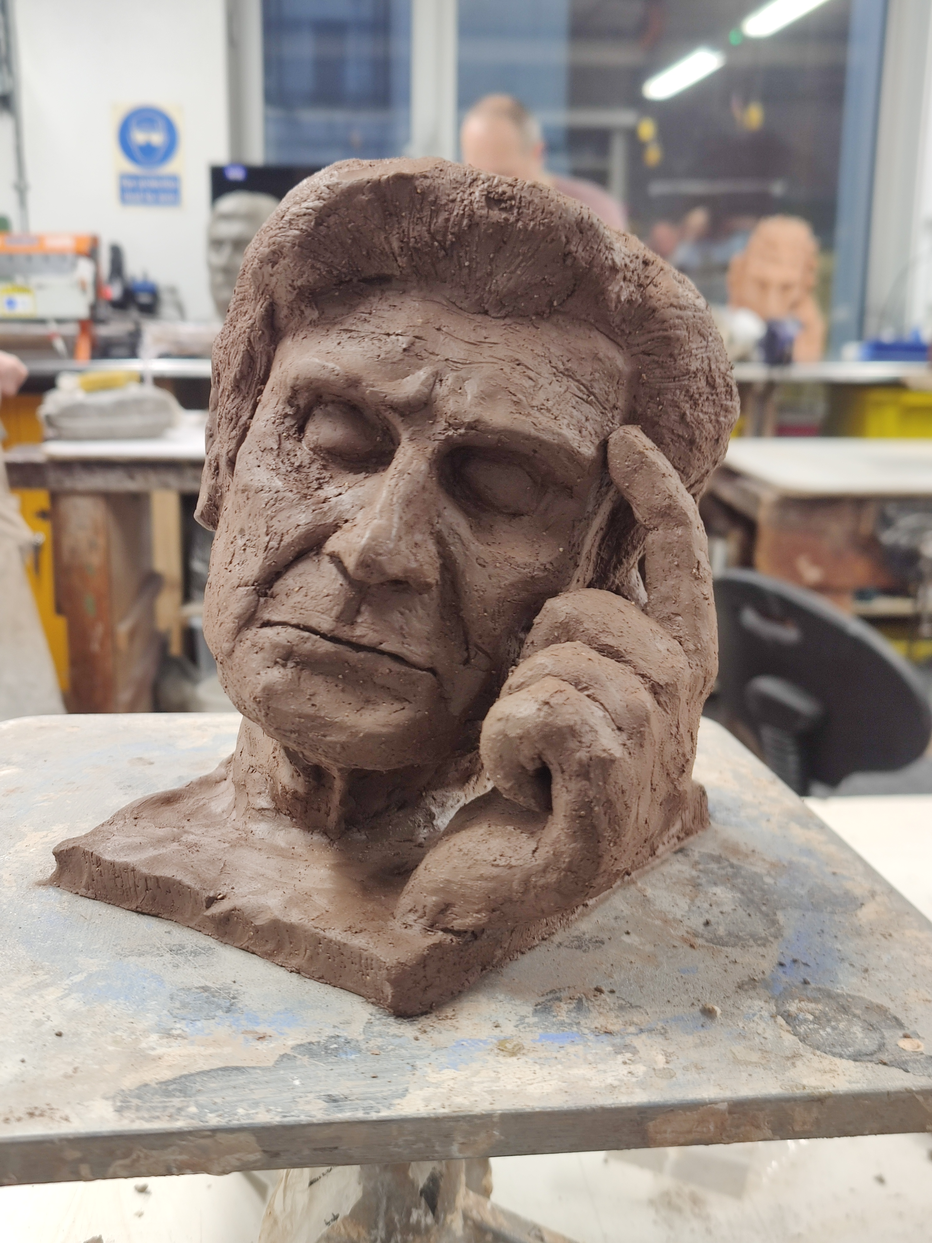Clay sculpture of a closed-eyed male head slightly tilted into a hand with index finger extended onto the left temple